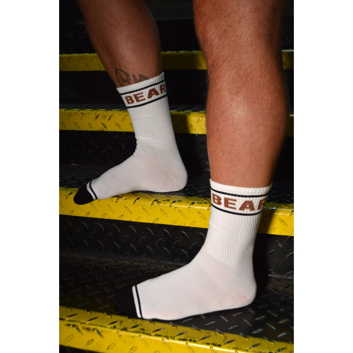 White socks with 'BEAR' branding on a person's feet, standing on a yellow and black textured surface.