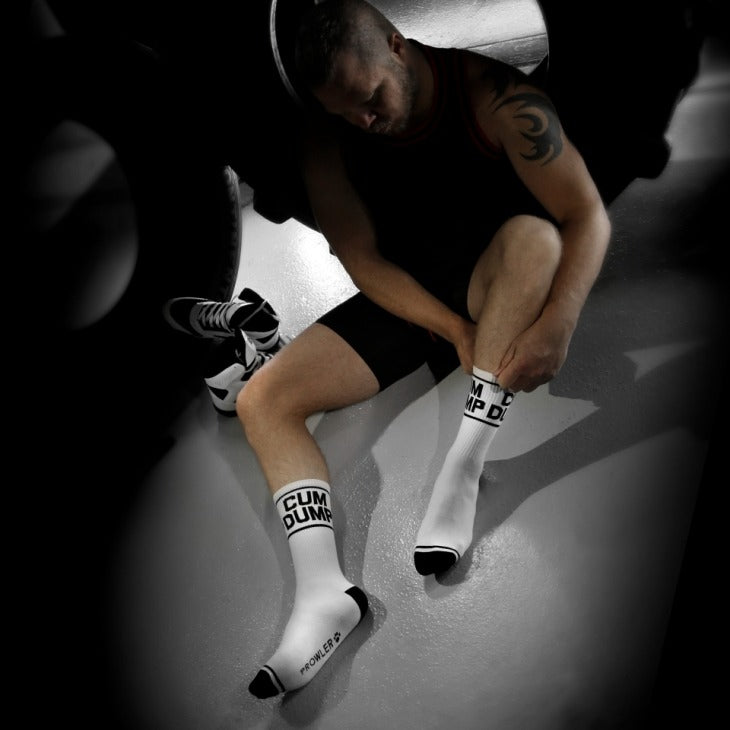 Man sitting on a concrete floor wearing white socks with text, surrounded by dark shadows.
