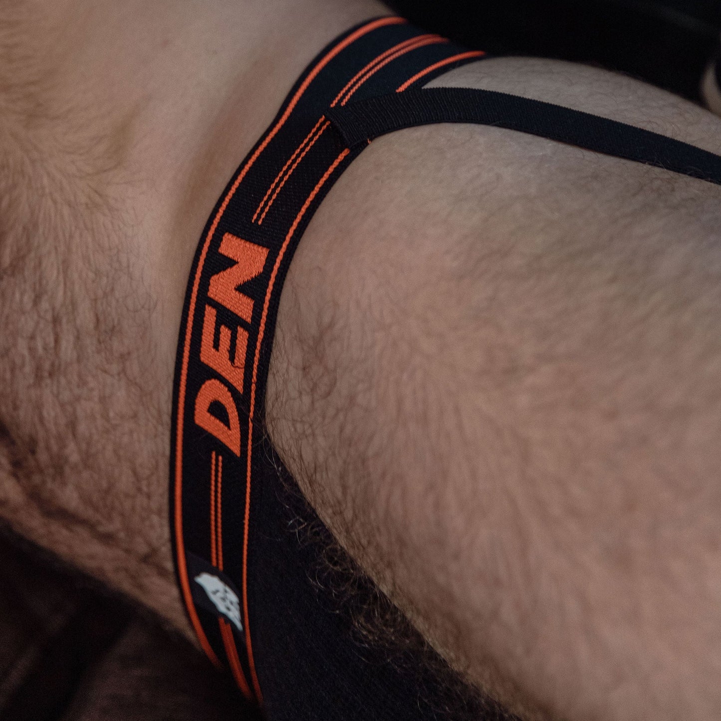 Close-up of a person wearing a black thong with orange 'DEN' branding.