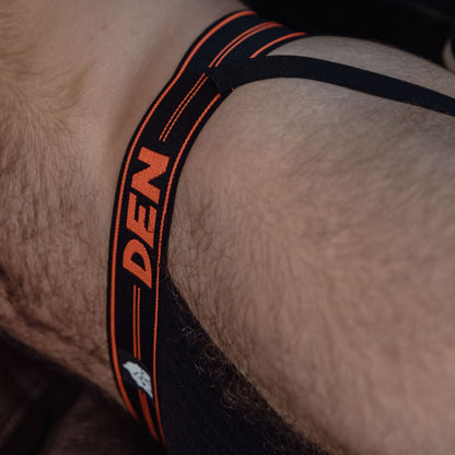 Close-up of a person wearing a black thong with orange 'DEN' branding.