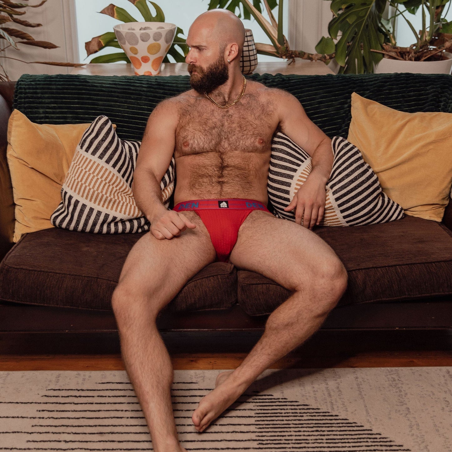 Man sitting on a couch wearing red underwear in a living room with plants.