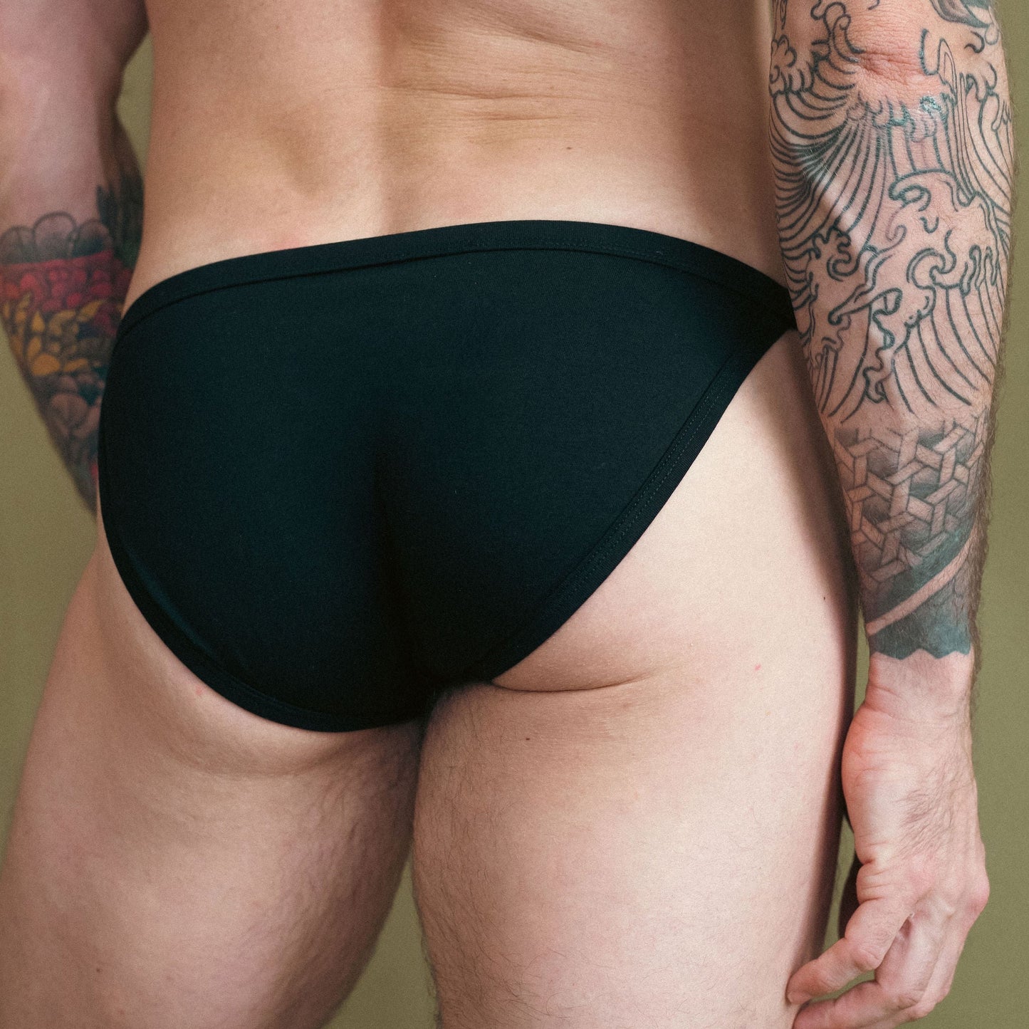 Person wearing black swim briefs with a tattooed arm on a plain background