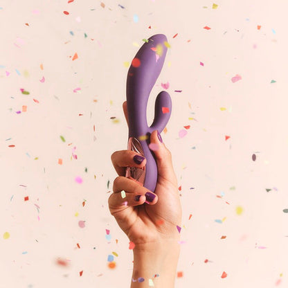 A hand holding a purple silicone personal massager against a light beige background with scattered confetti.