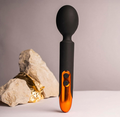 Black and orange handheld massager with a rock and gold nugget on a neutral background