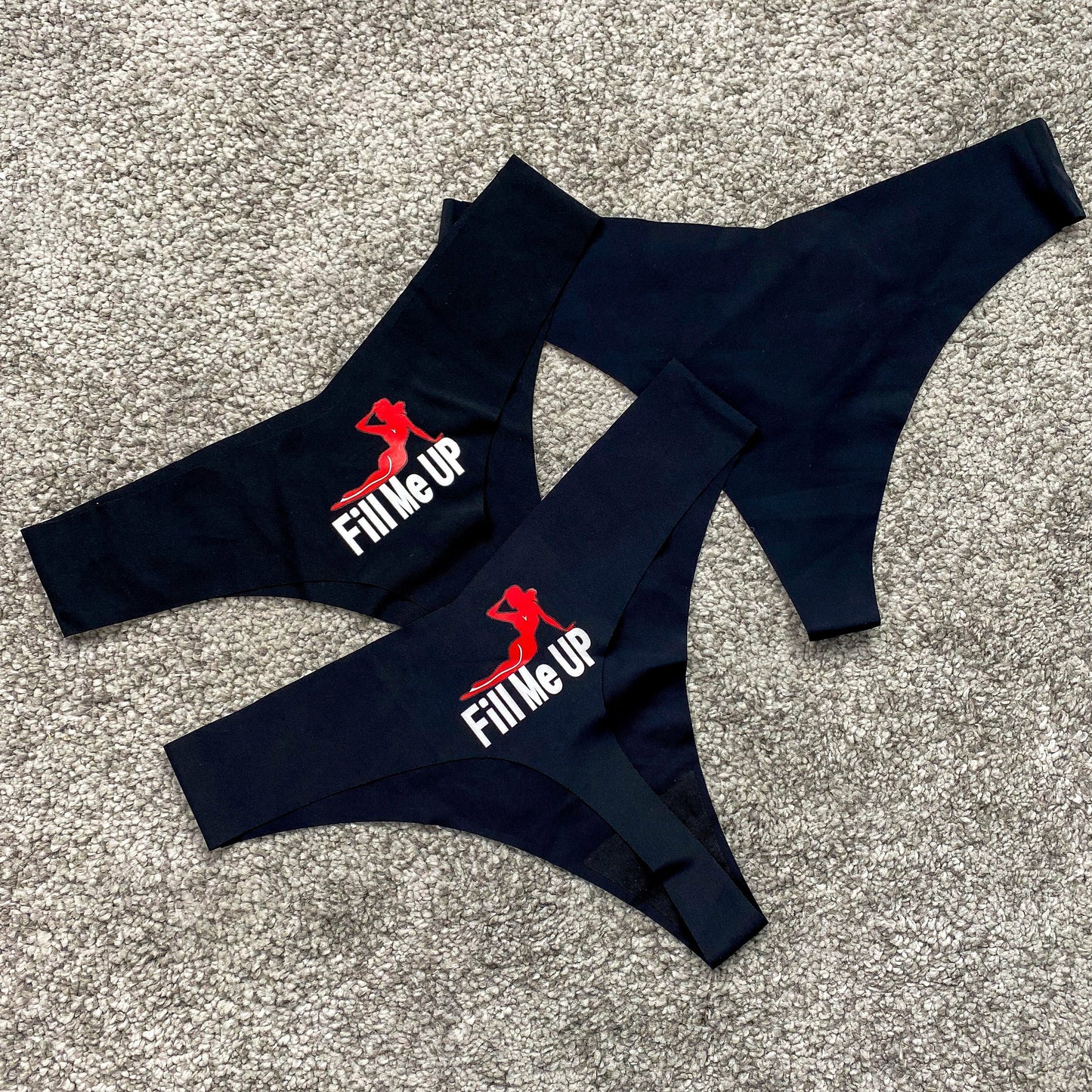 Two black thongs with 'Fill Me Up' logo on a textured surface