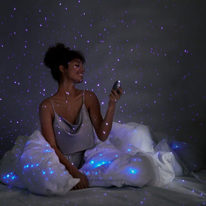 Woman sitting on a bed with starry light projector, creating a cosmic effect.