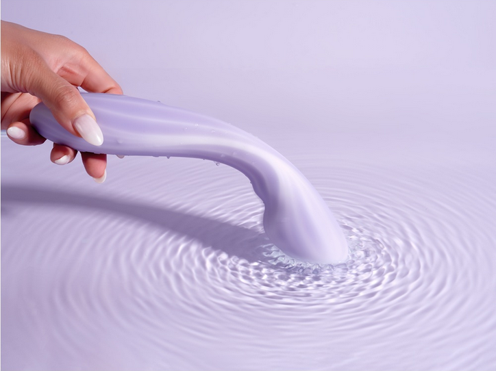 Hand holding a purple massager over water with ripples on a light purple background