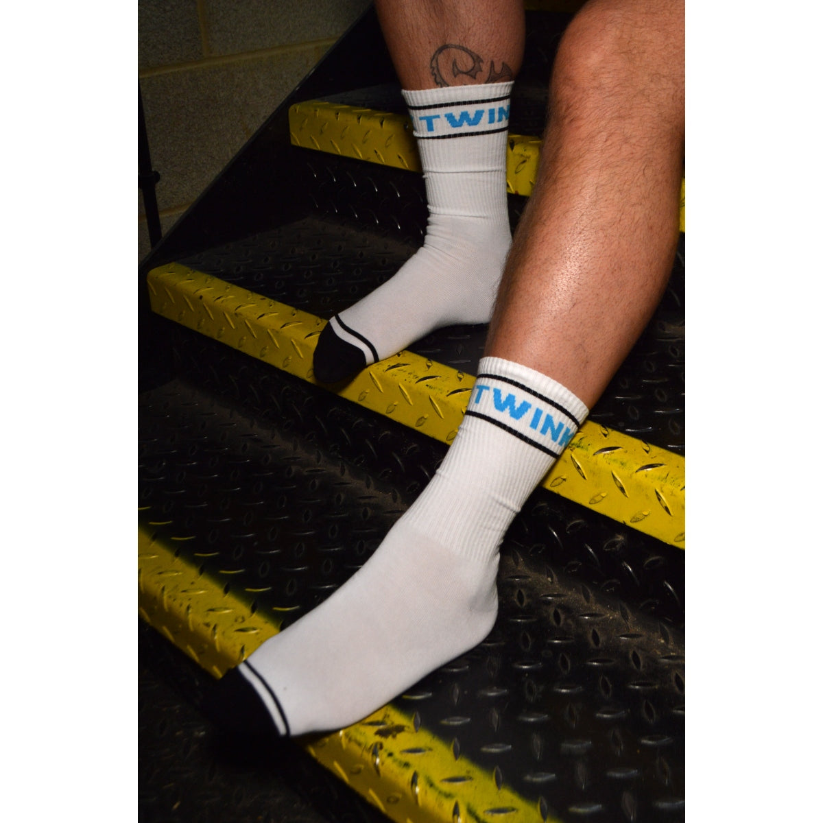White socks with black and blue branding on a person's feet, standing on a textured surface.