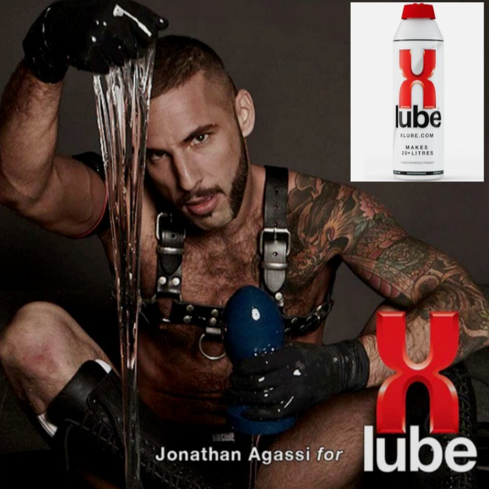 Man with tattoos holding a bottle of X Lube against a dark background