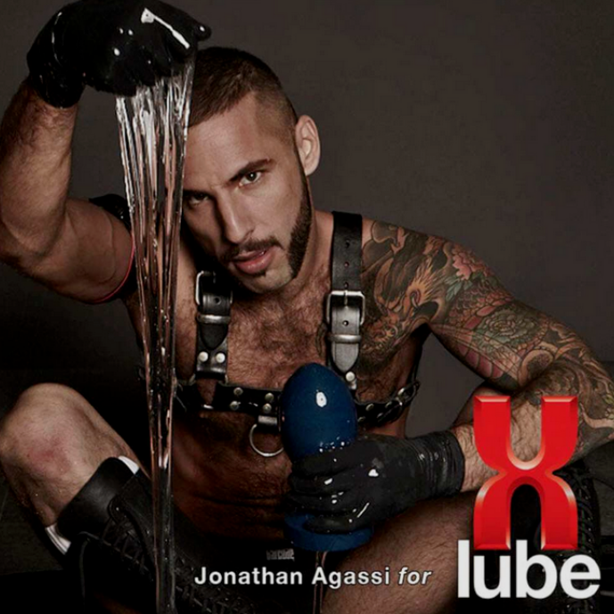 Man with tattoos holding a blue object, wearing a harness, with 'X Lube' branding.