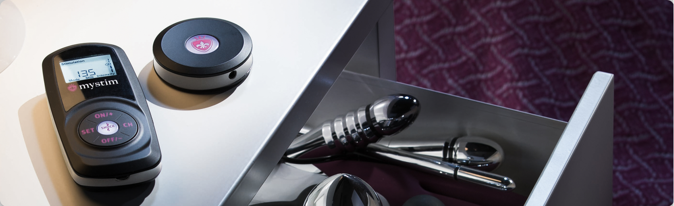 Two Mystim electronic devices on a table surface with sex toys in an open drawer below on a textured purple background